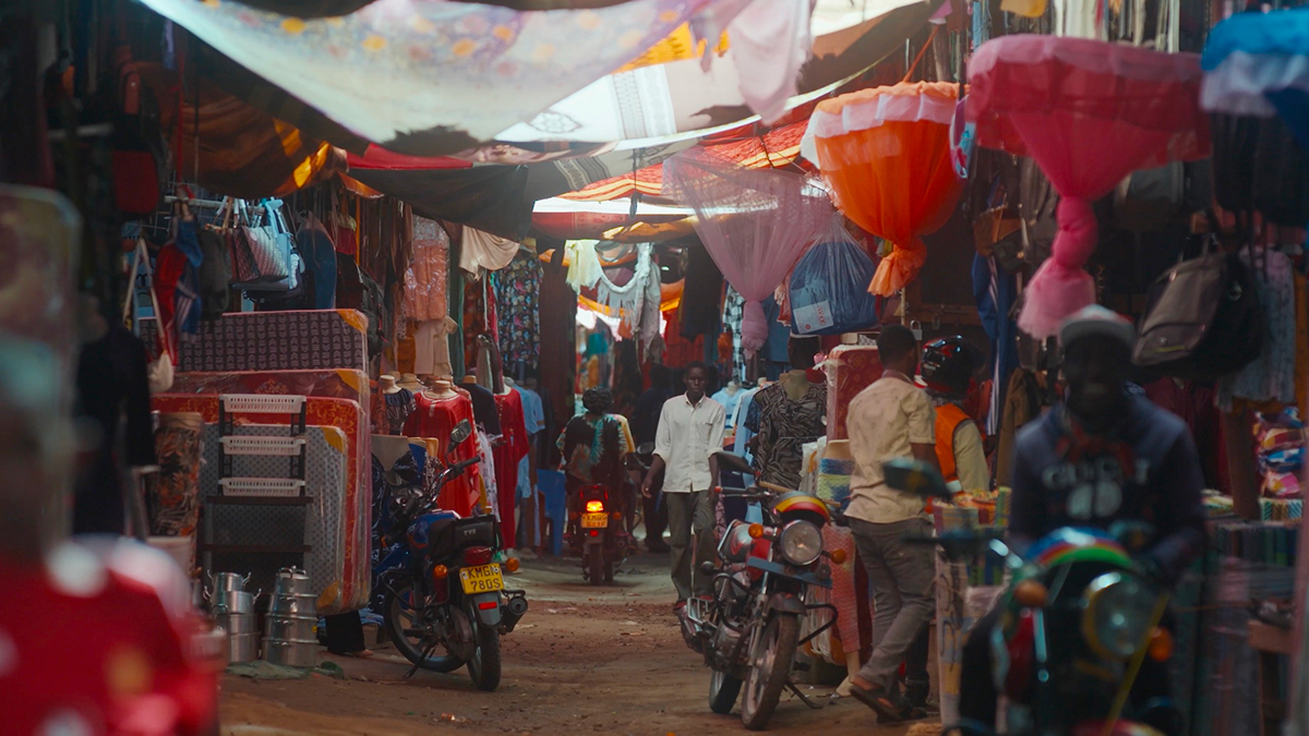 A bustling market scene with colorful fabric canopies overhead, motorbikes parked along the dirt pathway, and people walking and shopping among stalls selling clothes and household goods.